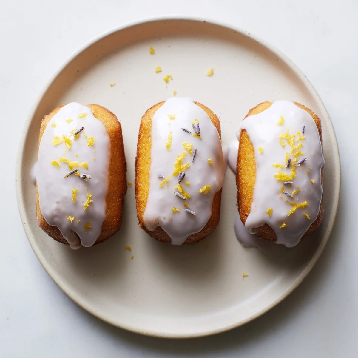Small floral glazed lemon cakes arranged on white plate with scattered dried lavender buds