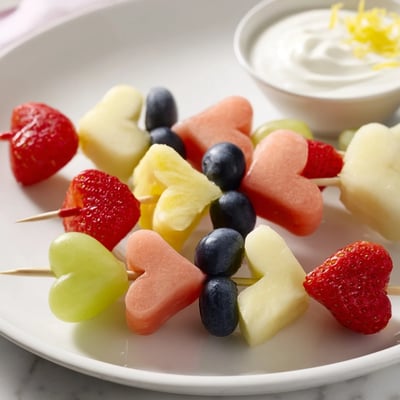 A hand dipping a Sweetheart Fruit Skewer with Yogurt Dip into the creamy honey glaze.
